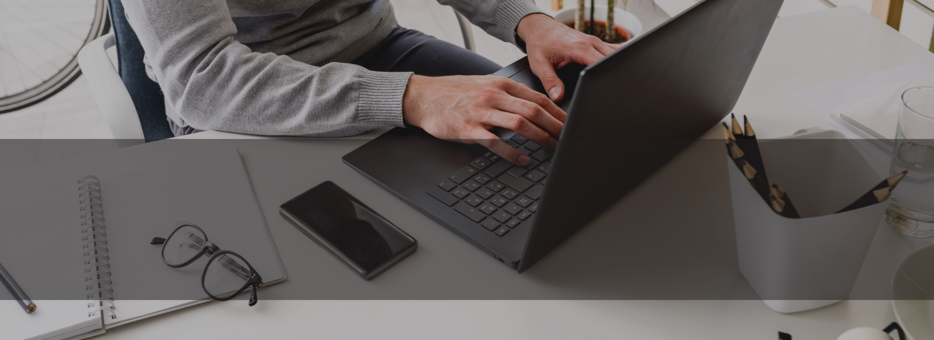 Homem de tom de pele branca com casaco na cor cinza sentado em uma cadeira de escritorio mexendo em um notebook na cor preta em cima de uma mesa de escritorio branca com um celular preto, um óculos e uma agenda também em cima da mesa