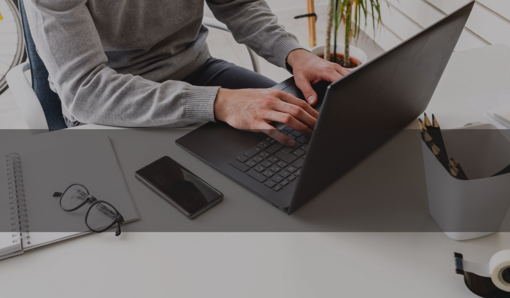 Homem de tom de pele branca com casaco na cor cinza sentado em uma cadeira de escritorio mexendo em um notebook na cor preta em cima de uma mesa de escritorio branca com um celular preto, um óculos e uma agenda também em cima da mesa